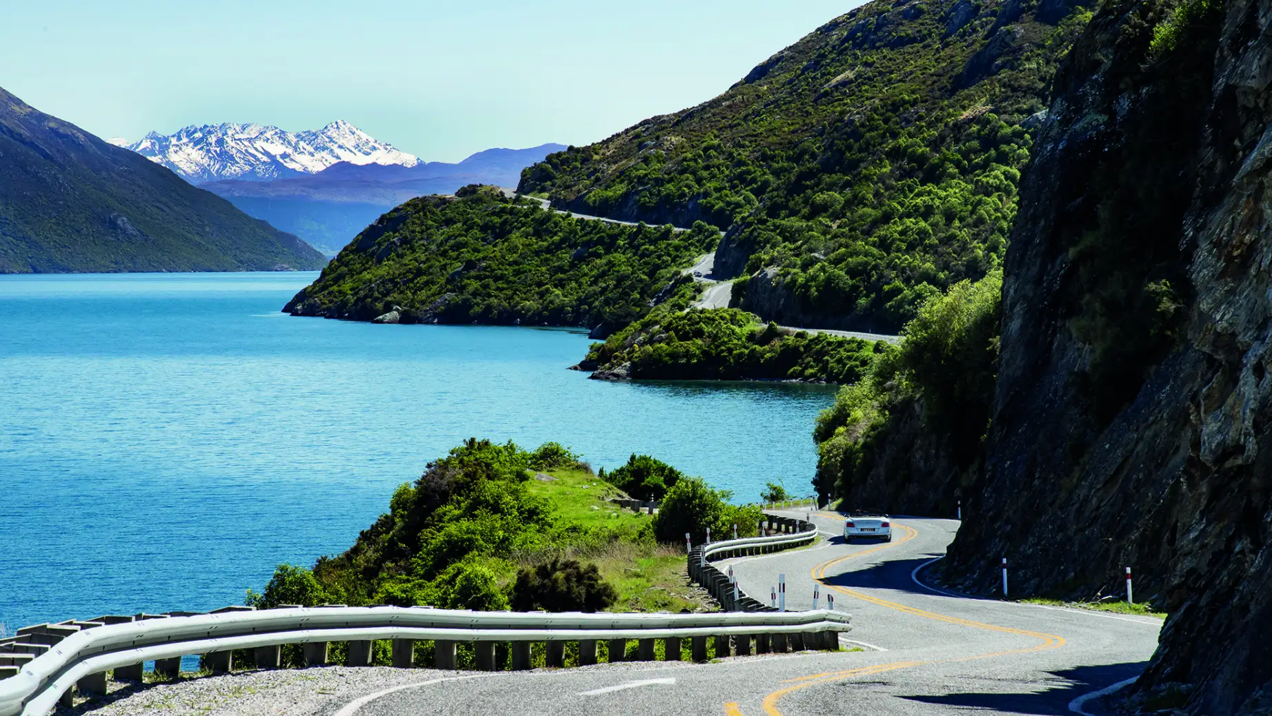 Kurvenreiche Straße in Neuseeland, mit Auto zwischen blauem Gewässer grün bewachsener Felslandschaft und schneebedecktem Gebirge im Hintergrund