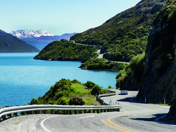 Kurvenreiche Straße in Neuseeland, mit Auto zwischen blauem Gewässer grün bewachsener Felslandschaft und schneebedecktem Gebirge im Hintergrund