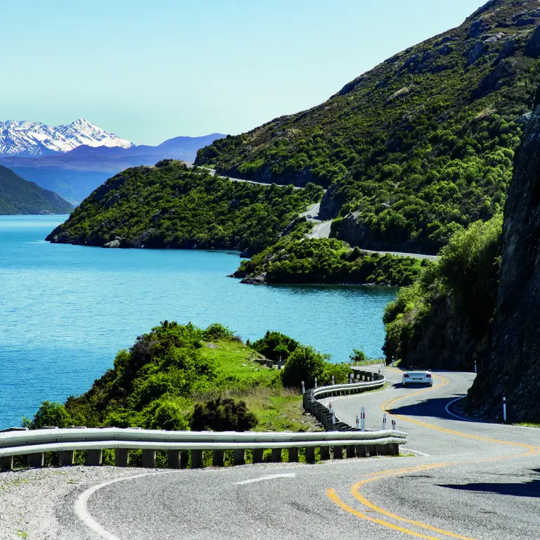 Kurvenreiche Straße in Neuseeland, mit Auto zwischen blauem Gewässer grün bewachsener Felslandschaft und schneebedecktem Gebirge im Hintergrund
