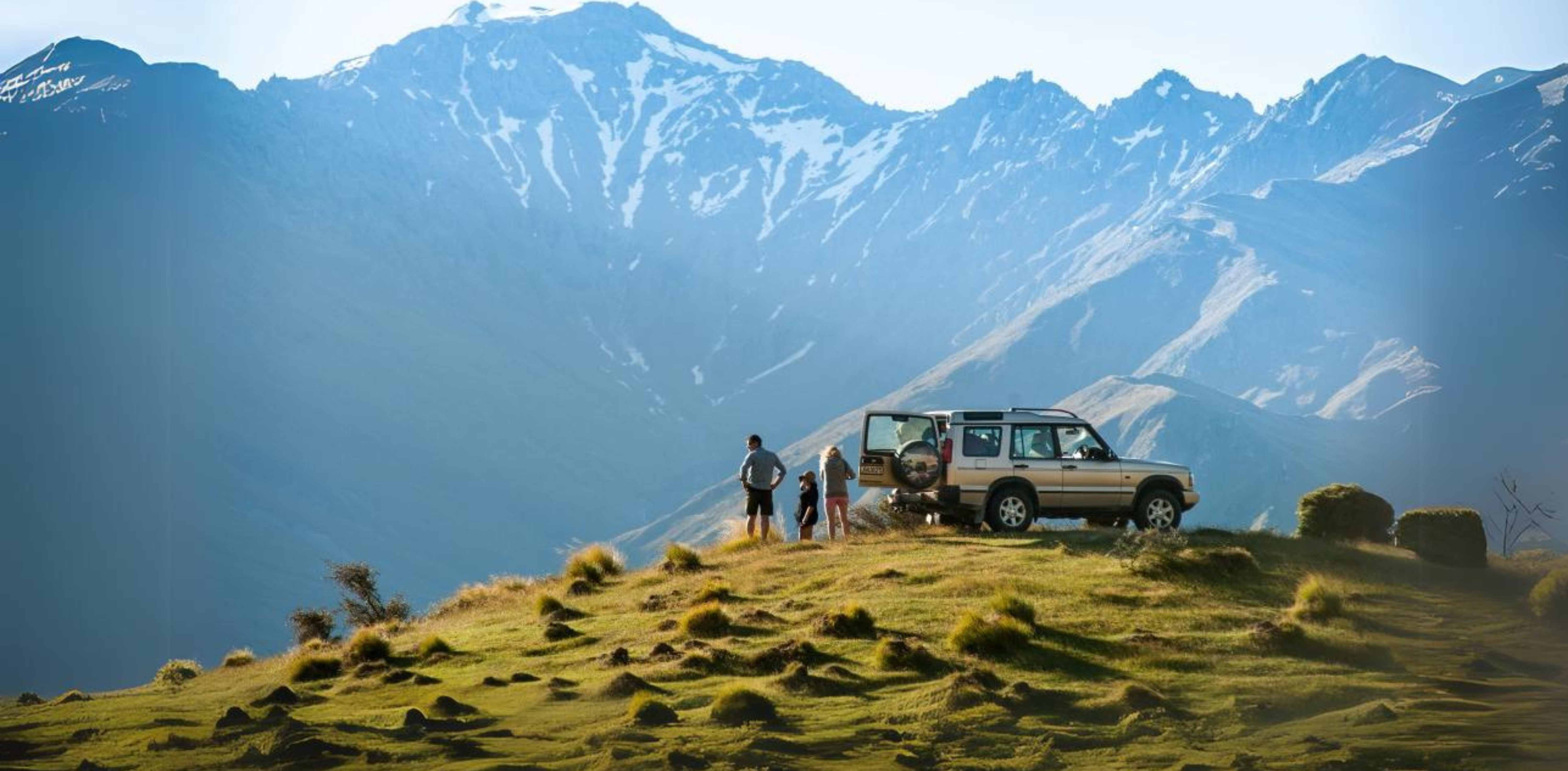 Eine Familie steht neben einem Auto am Rande eines Hügels mit Blick auf die Berge in Neuseeland