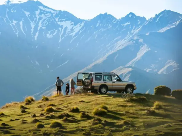 A family stand next to a car on the side of a hill overlooking the mountains in New Zealand