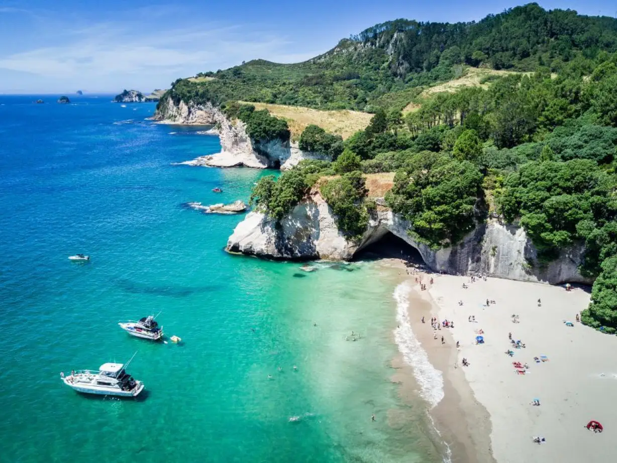 Aerial view of Cathedral Cove, New Zealand with boats moored in sea and people relaxing on the beach