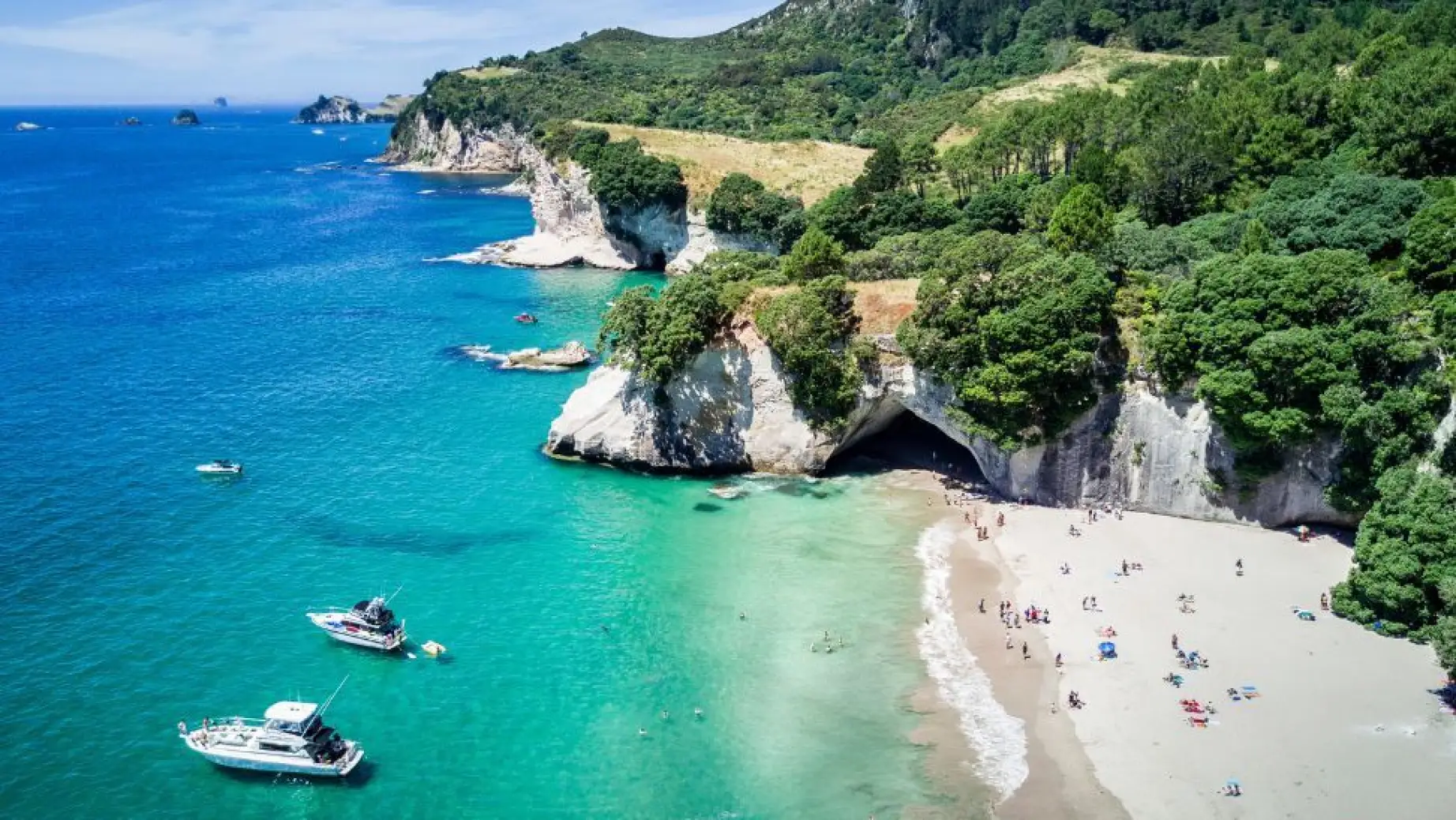 Luftaufnahme von Cathedral Cove, Neuseeland, mit Booten, die im Meer vor Anker liegen, und Menschen, die sich am Strand entspannen.