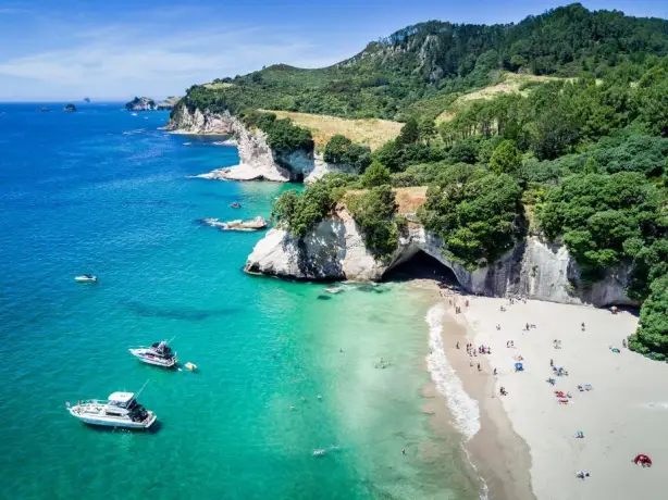 Luftaufnahme von Cathedral Cove, Neuseeland, mit Booten, die im Meer vor Anker liegen, und Menschen, die sich am Strand entspannen.