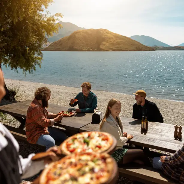 Een groep mensen geniet van drankjes en pizza aan picknicktafels op een terras