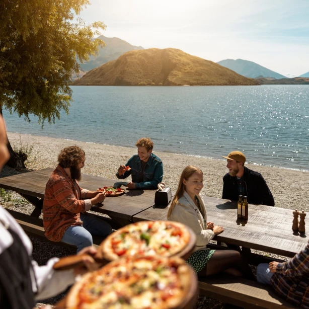 Een groep mensen geniet van drankjes en pizza aan picknicktafels op een terras