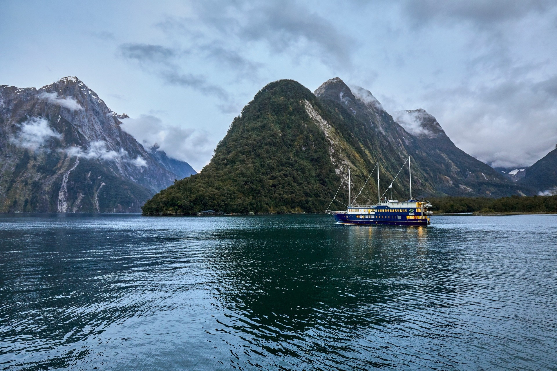 A cruise ship glides through Milford Sound in Fiordland National Park, surrounded by steep, forested mountains and dark, calm water beneath a cloudy sky.
