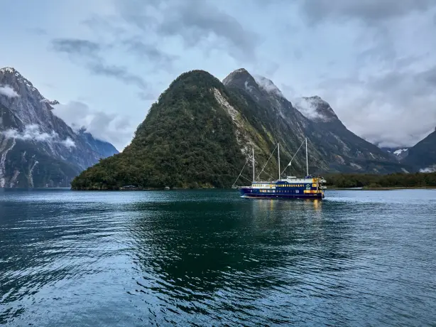 A cruise ship glides through Milford Sound in Fiordland National Park, surrounded by steep, forested mountains and dark, calm water beneath a cloudy sky.