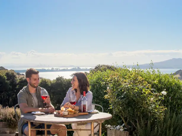 A couple drinking wine and eating cheese against a backdrop of mountains and sea on Waikheke Island, New Zealand