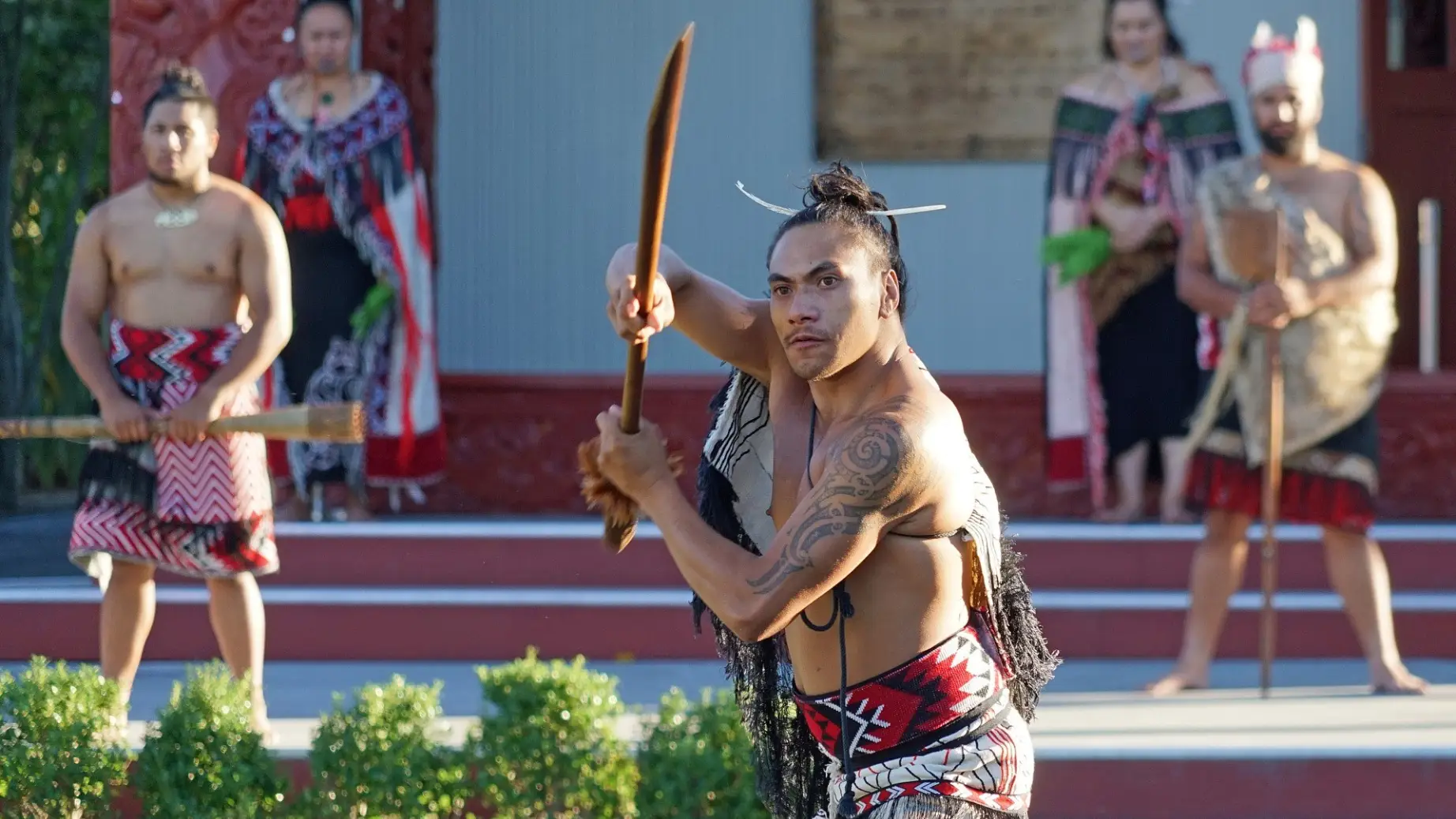 Eine Gruppe Māori-Männer in traditioneller Kleidung hält hölzerne Waffen (Taiaha)
