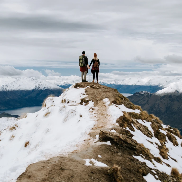 Couple stand at mountain summit looking over mountain peaks and fjords