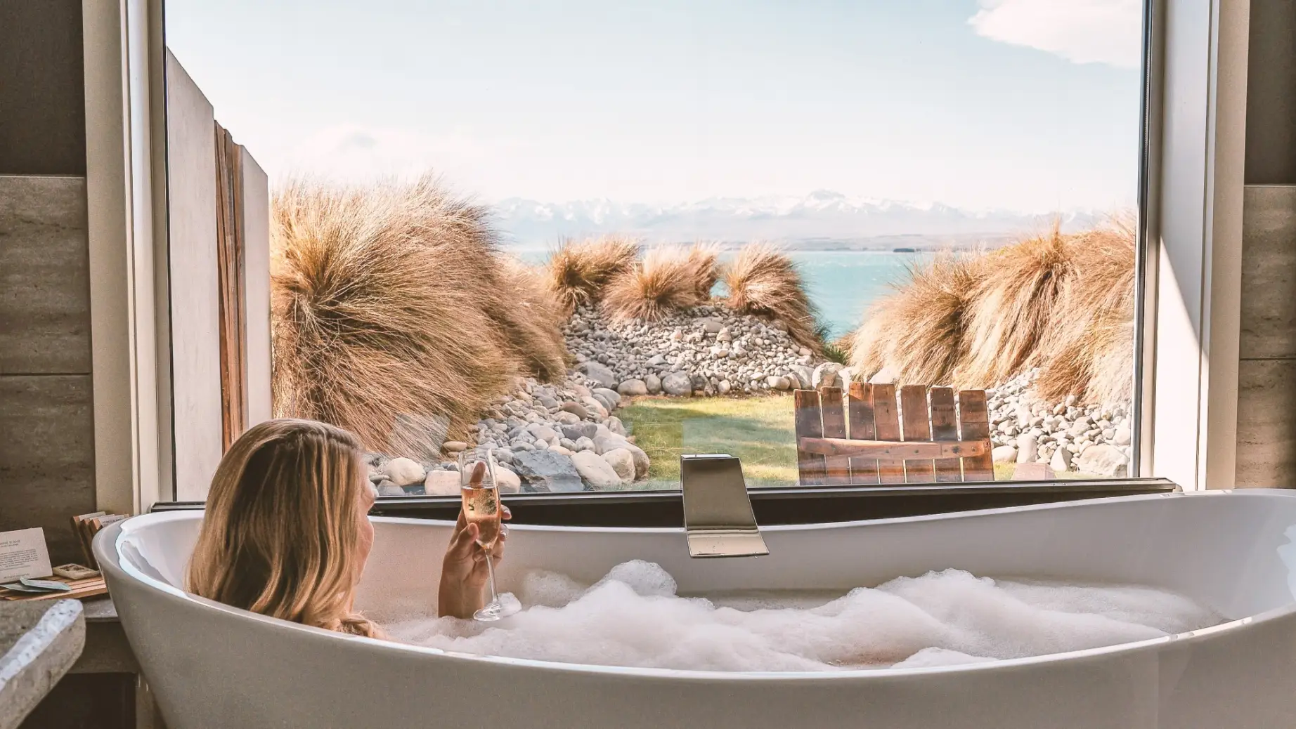 Frau sitzt mit Glas Prosecco in der Hand in einer schaumgefüllten Badewanne mit Panoramablick über den See