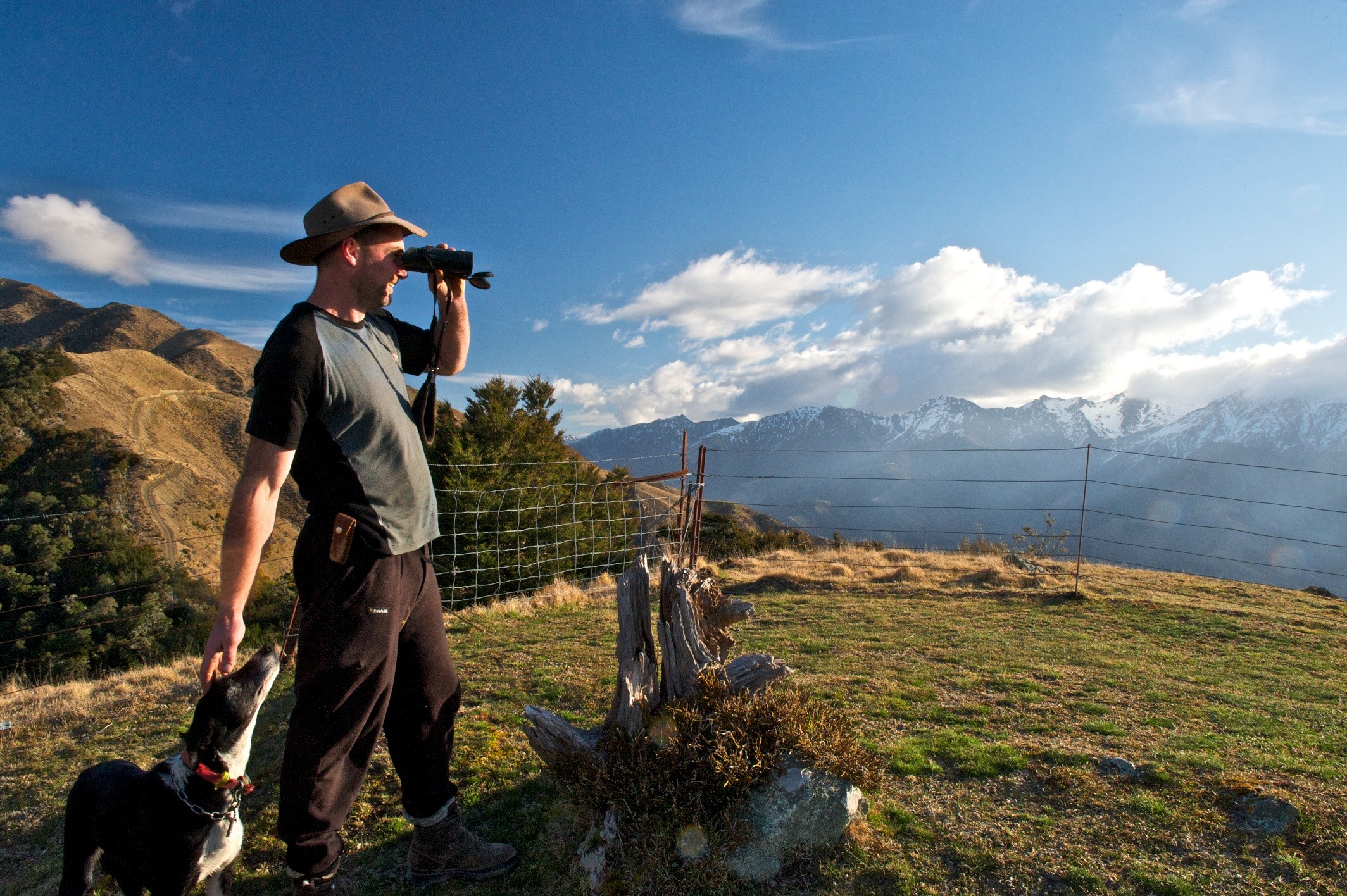 Mann mit Hut und einem Hund schaut durch ein Fernglas auf Berge in der Ferne