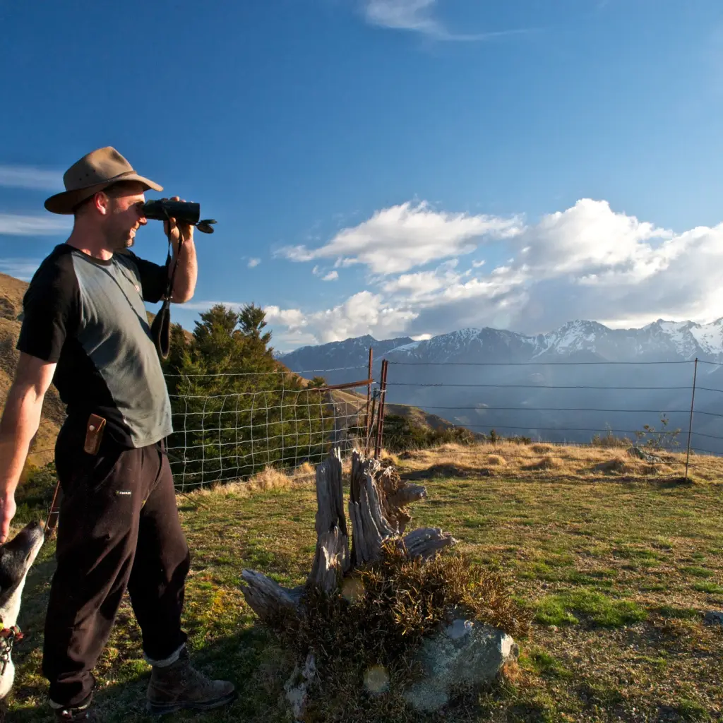 Mann mit Hut und einem Hund schaut durch ein Fernglas auf Berge in der Ferne