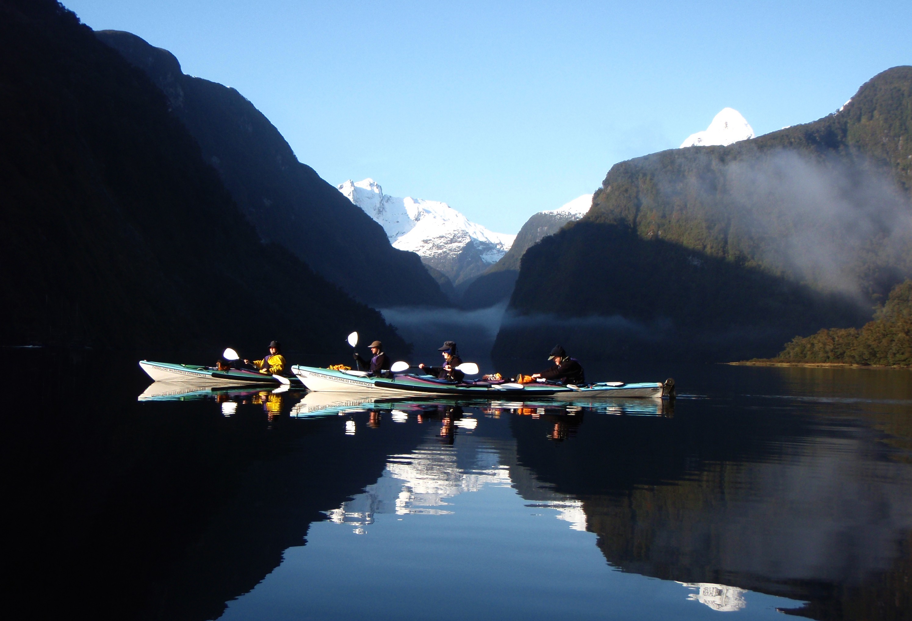 A group kayaking among green cliffs and snowy peaks in Doubtful Sound, Fiordland National Park