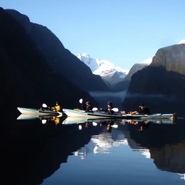 A group kayaking among green cliffs and snowy peaks in Doubtful Sound, Fiordland National Park