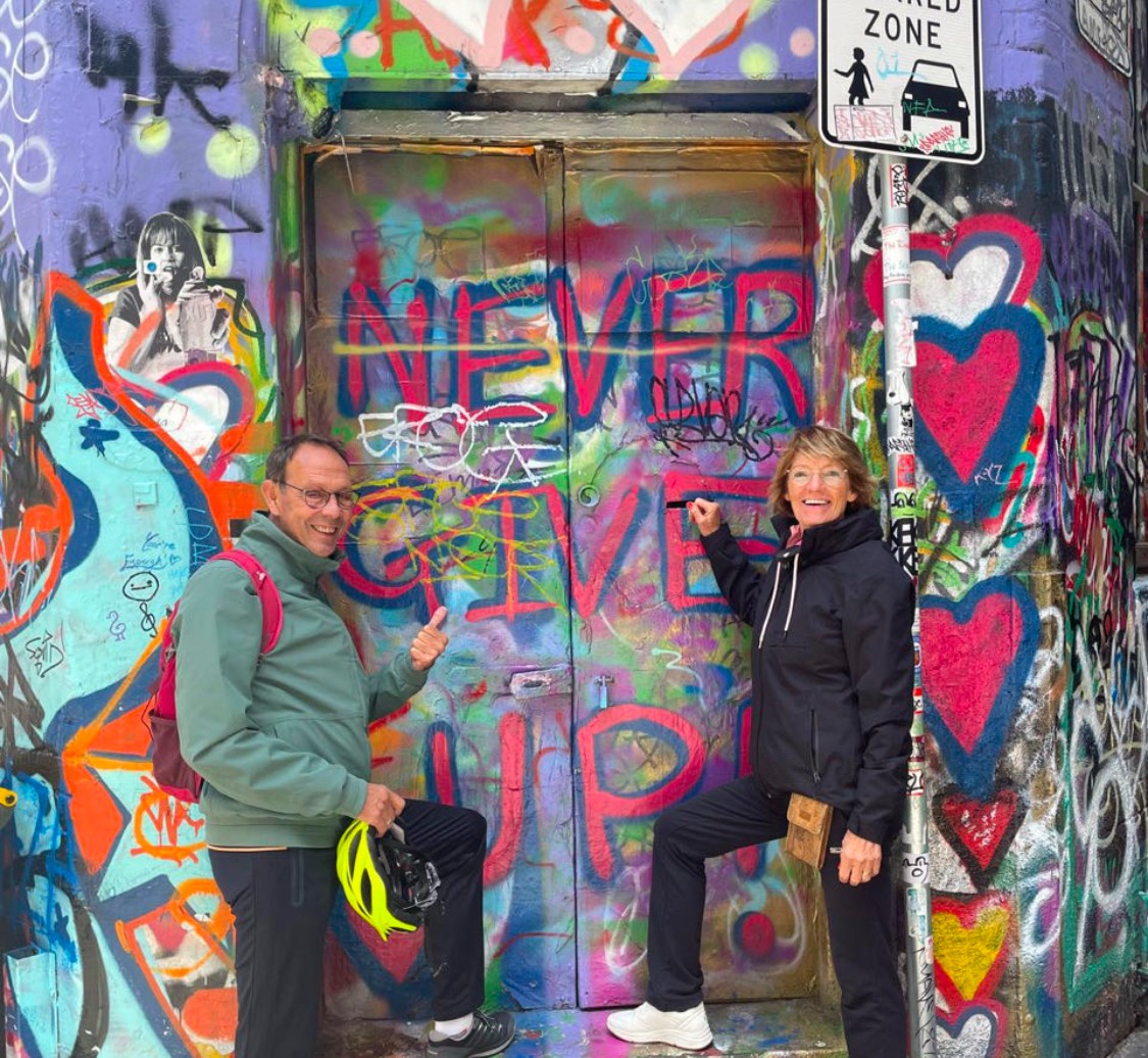 Paulien en Norbert poseren bij een graffiti muur met "Never Give Up" erop
