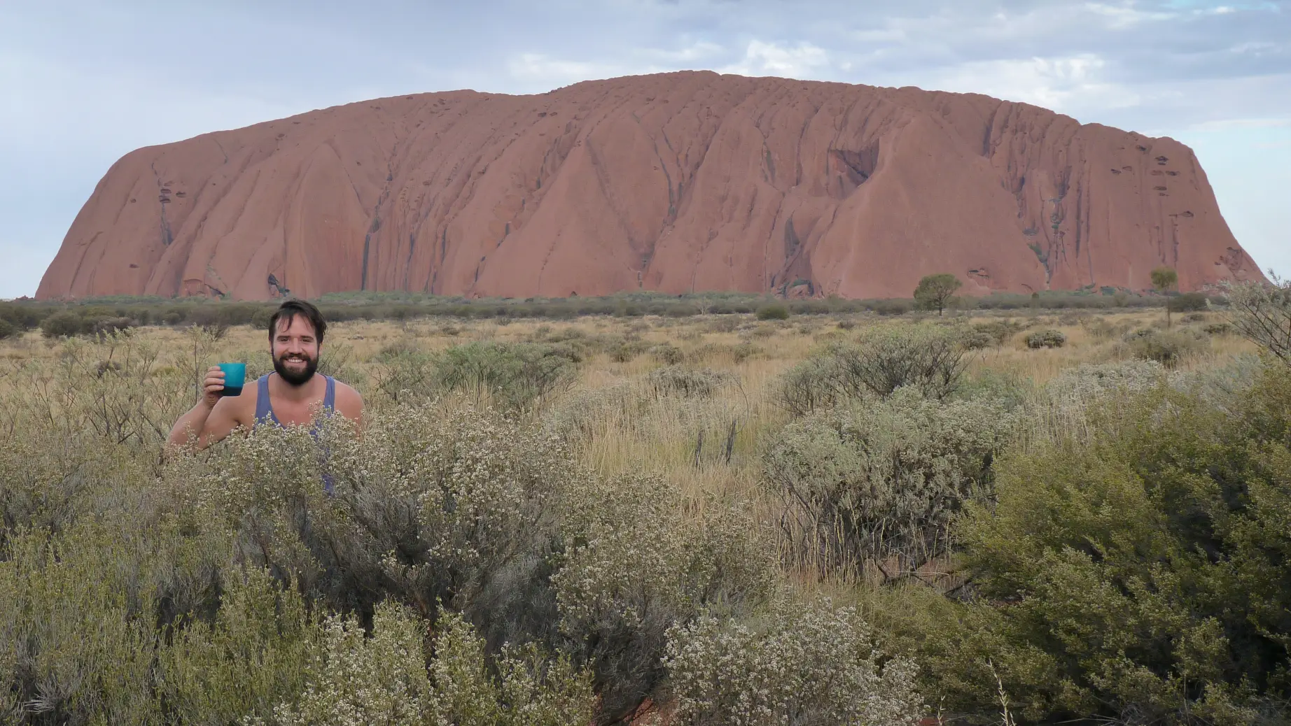 A man stands behind a bush with a glass of drink in his hand with Ayers Rock/Uluru in the background in Australia's Red Centre