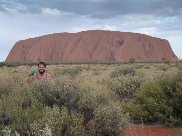 A man stands behind a bush with a glass of drink in his hand with Ayers Rock/Uluru in the background in Australia's Red Centre