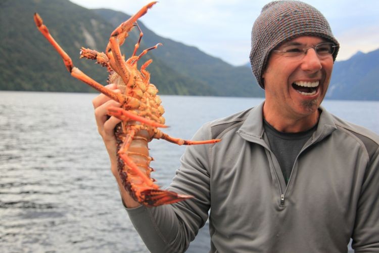A man wearing a beanie hat picks up a crayfish while smiling on a boat in Doubtful Sound, Fiordland National Park, New Zealand