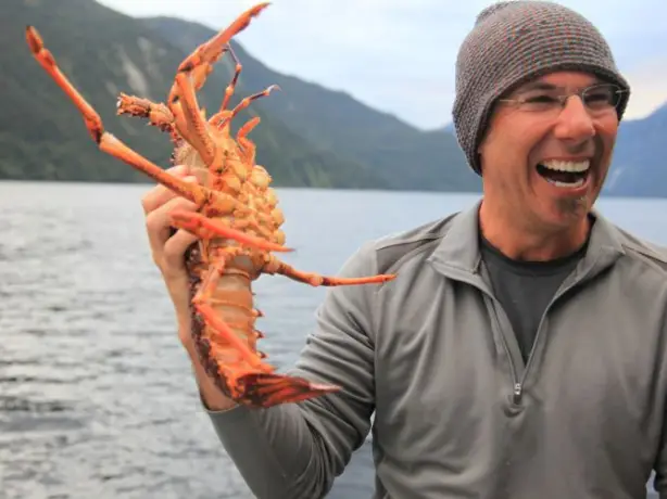 A man wearing a beanie hat picks up a crayfish while smiling on a boat in Doubtful Sound, Fiordland National Park, New Zealand