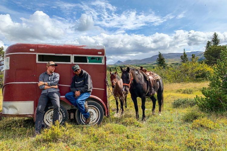 Zwei Männer lehnen an einem roten Anhänger, an dem zwei Pferde angebunden sind, vor dem Hintergrund sanfter Hügel und blauem Himmel in Montana