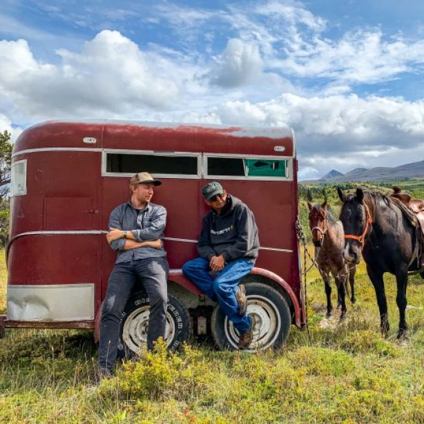 Zwei Männer lehnen an einem roten Anhänger, an dem zwei Pferde angebunden sind, vor dem Hintergrund sanfter Hügel und blauem Himmel in Montana