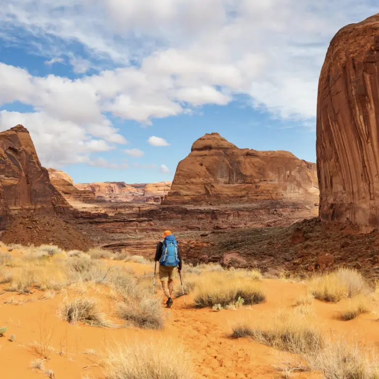 Een solo-wandelaar met een blauwe rugzak en wandelstokken die door een woestijnlandschap van rood zand trekt, te midden van torenhoge zandstenen rotsformaties in Monument Valley, Utah, VS