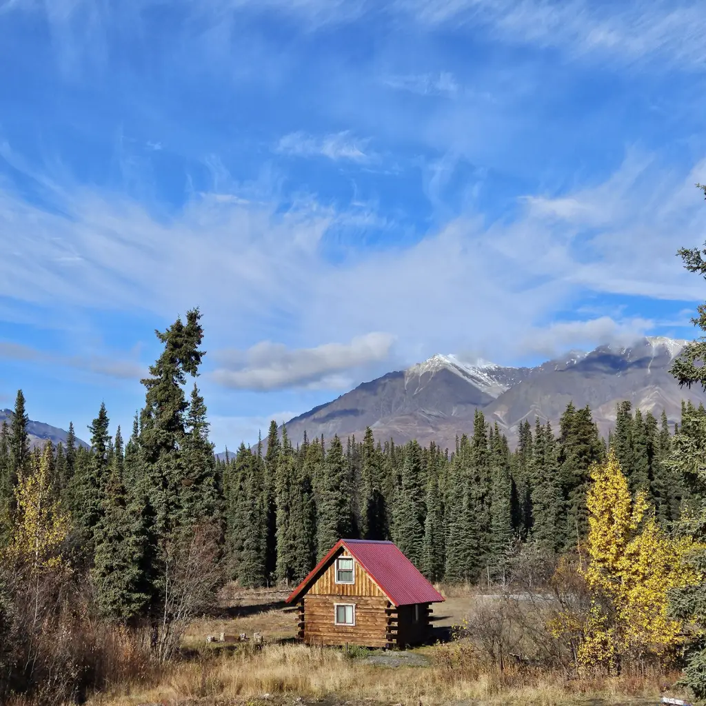 Eenzame blokhut met een rood dak, genesteld tussen sparrenbomen en herfstbladeren in Wrangell-St. Elias National Park, Alaska, VS, met besneeuwde bergtoppen onder een dramatische blauwe lucht.