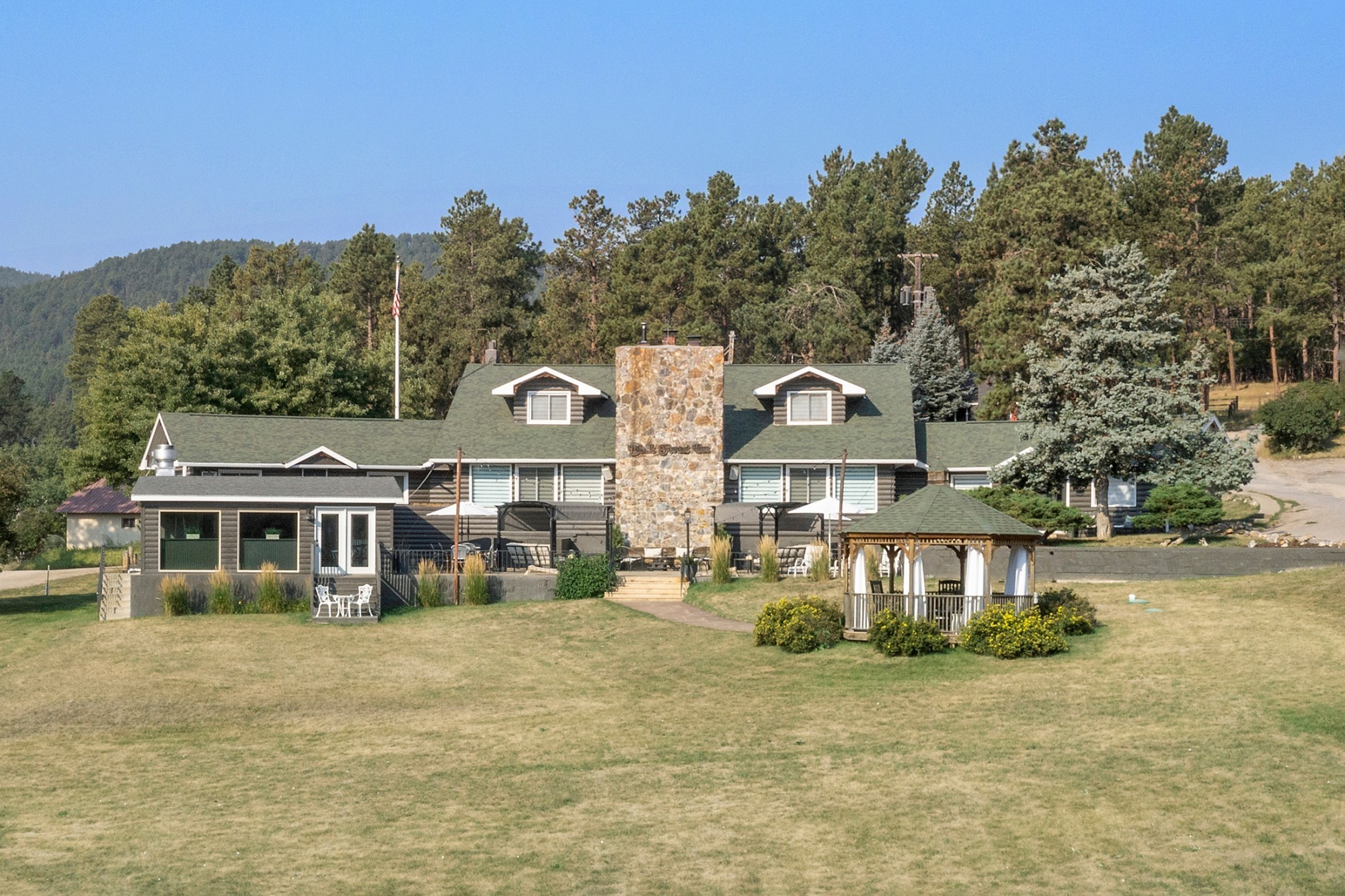 Rustikales Blockhaus-Inn mit hohem Steinkamin und Pavillon auf einer offenen Rasenfläche, umgeben von kiefernbedeckten Hügeln in der Black Hills Region von South Dakota, USA