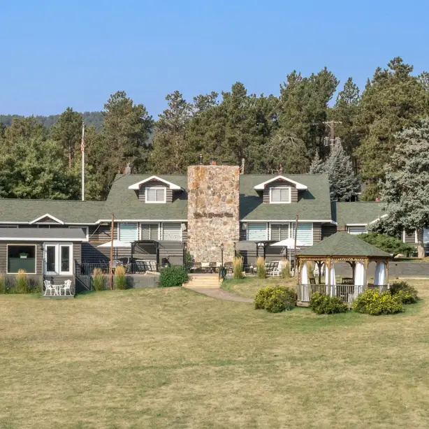 Rustikales Blockhaus-Inn mit hohem Steinkamin und Pavillon auf einer offenen Rasenfläche, umgeben von kiefernbedeckten Hügeln in der Black Hills Region von South Dakota, USA