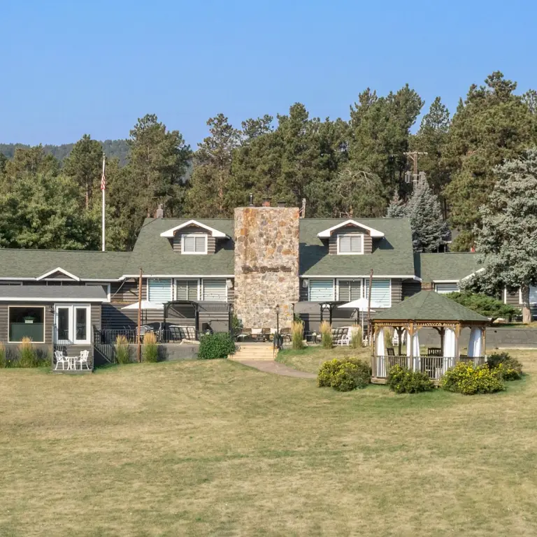 Rustikales Blockhaus-Inn mit hohem Steinkamin und Pavillon auf einer offenen Rasenfläche, umgeben von kiefernbedeckten Hügeln in der Black Hills Region von South Dakota, USA