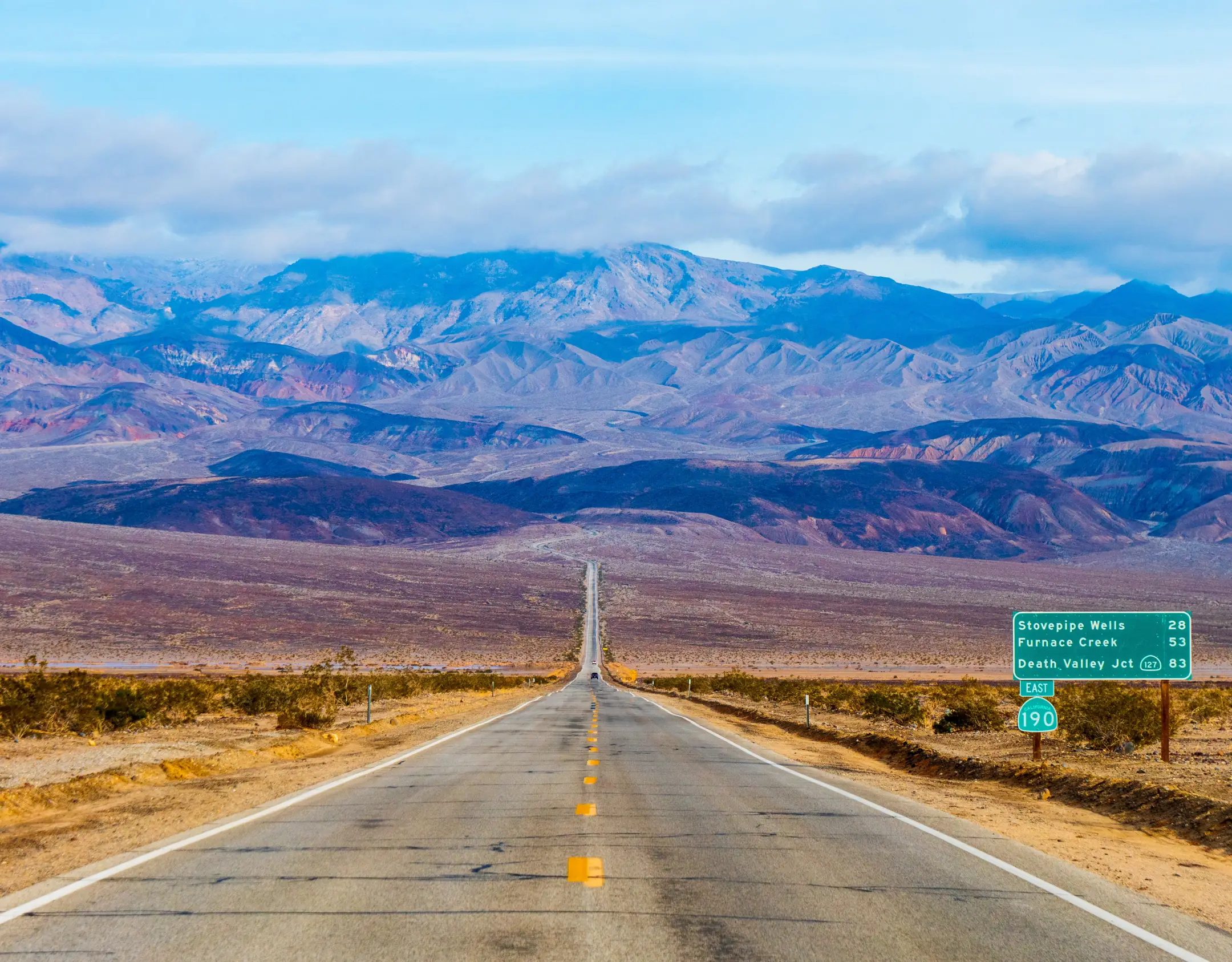 Eine lange, gerade Straße, die sich durch die kalifornische Mojave-Wüste in Richtung der Berge des Death Valley erstreckt, mit Wüstenebenen und fernen Bergrücken unter einem hellen Himmel.