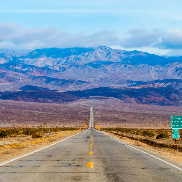 Eine lange, gerade Straße, die sich durch die kalifornische Mojave-Wüste in Richtung der Berge des Death Valley erstreckt, mit Wüstenebenen und fernen Bergrücken unter einem hellen Himmel.