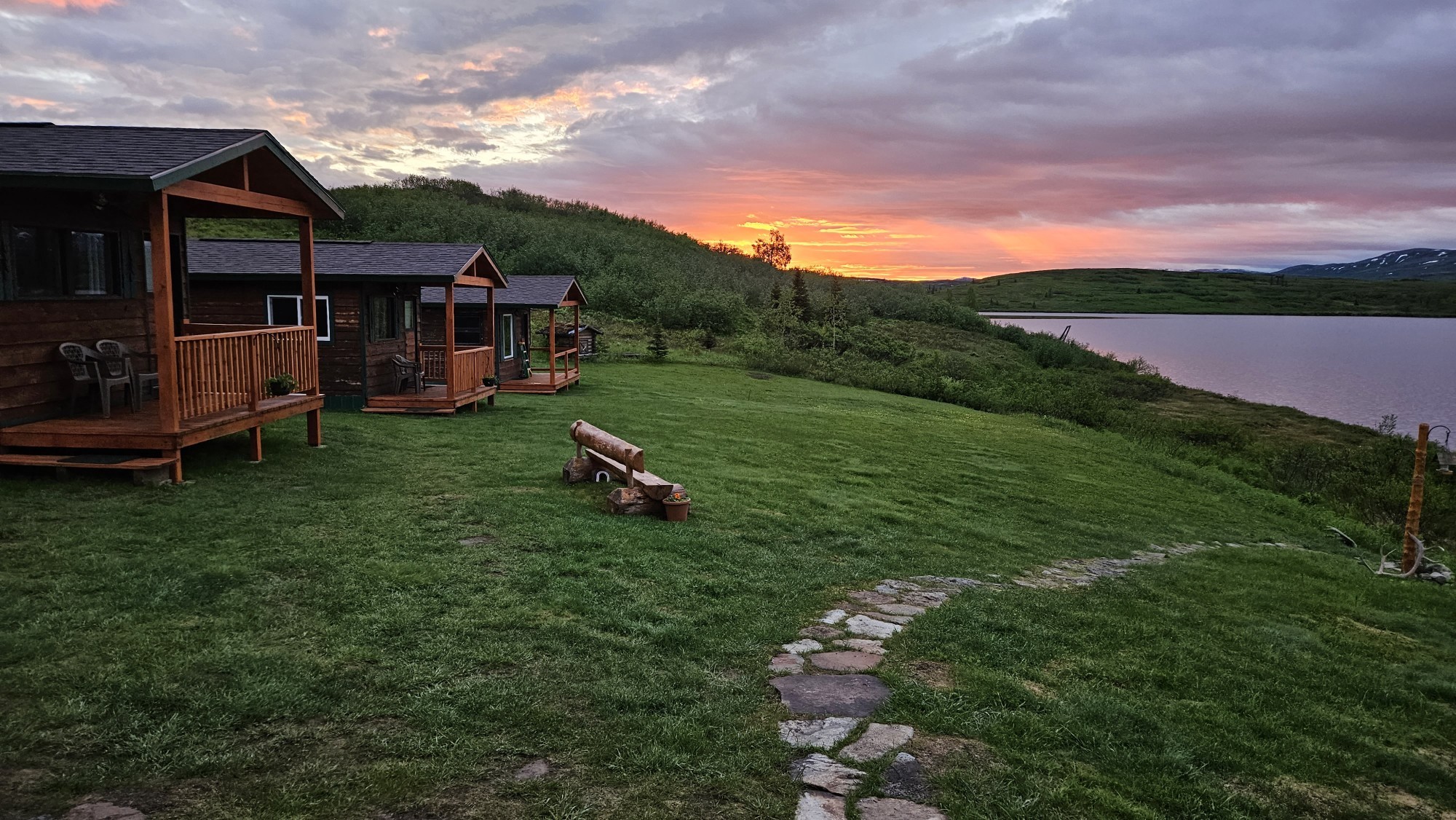 Wildnis-Lodge-Hütten mit überdachten Veranden auf einem grünen Hang mit Blick auf einen See in Alaska, USA, unter einem dramatischen feurig roten und orangefarbenen Mitternachtssonnenhimmel