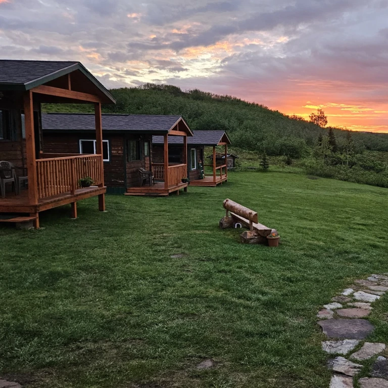 Wildnis-Lodge-Hütten mit überdachten Veranden auf einem grünen Hang mit Blick auf einen See in Alaska, USA, unter einem dramatischen feurig roten und orangefarbenen Mitternachtssonnenhimmel