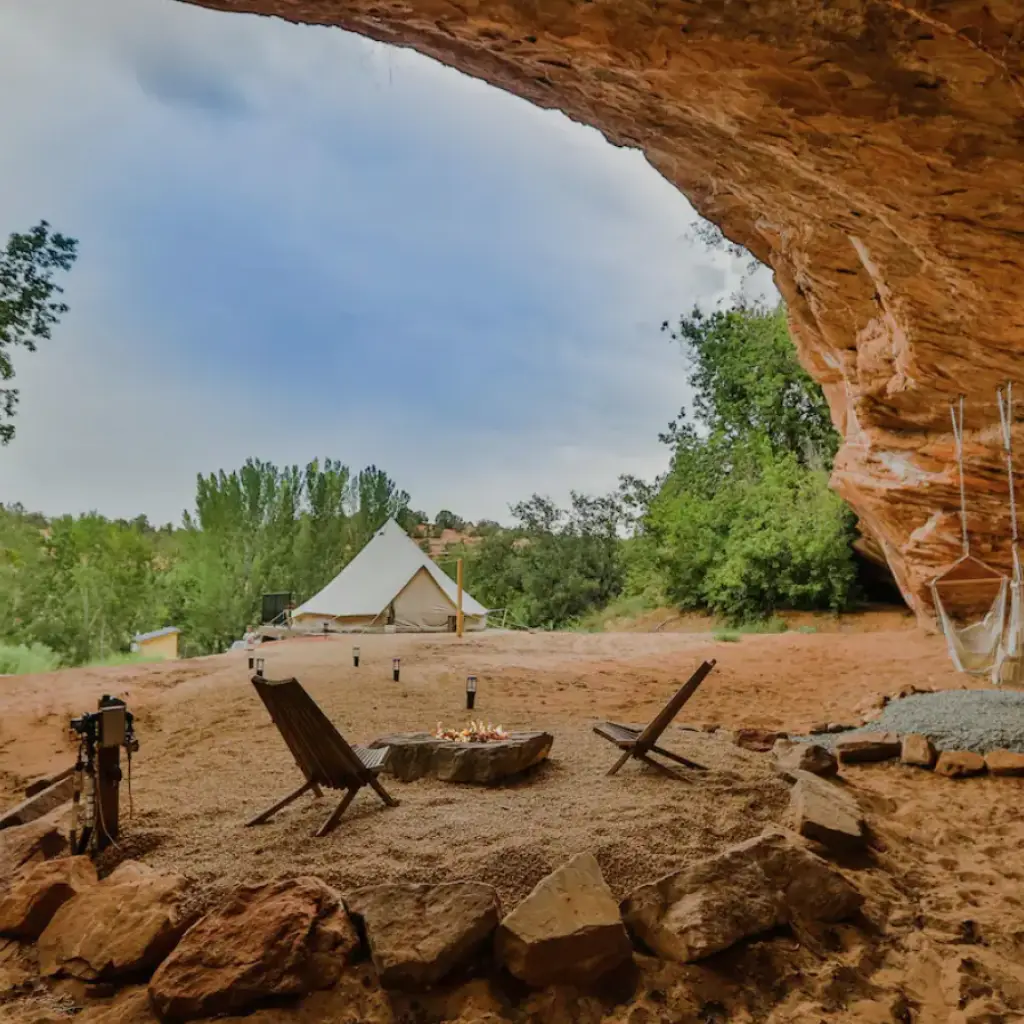 Glamping-Platz unter einem natürlichen Sandstein-Felsüberhang in Utah, USA, mit einem Leinwand-Glockenzelt, Lagerfeuer und Holzstühlen in einer roten Wüstenschlucht