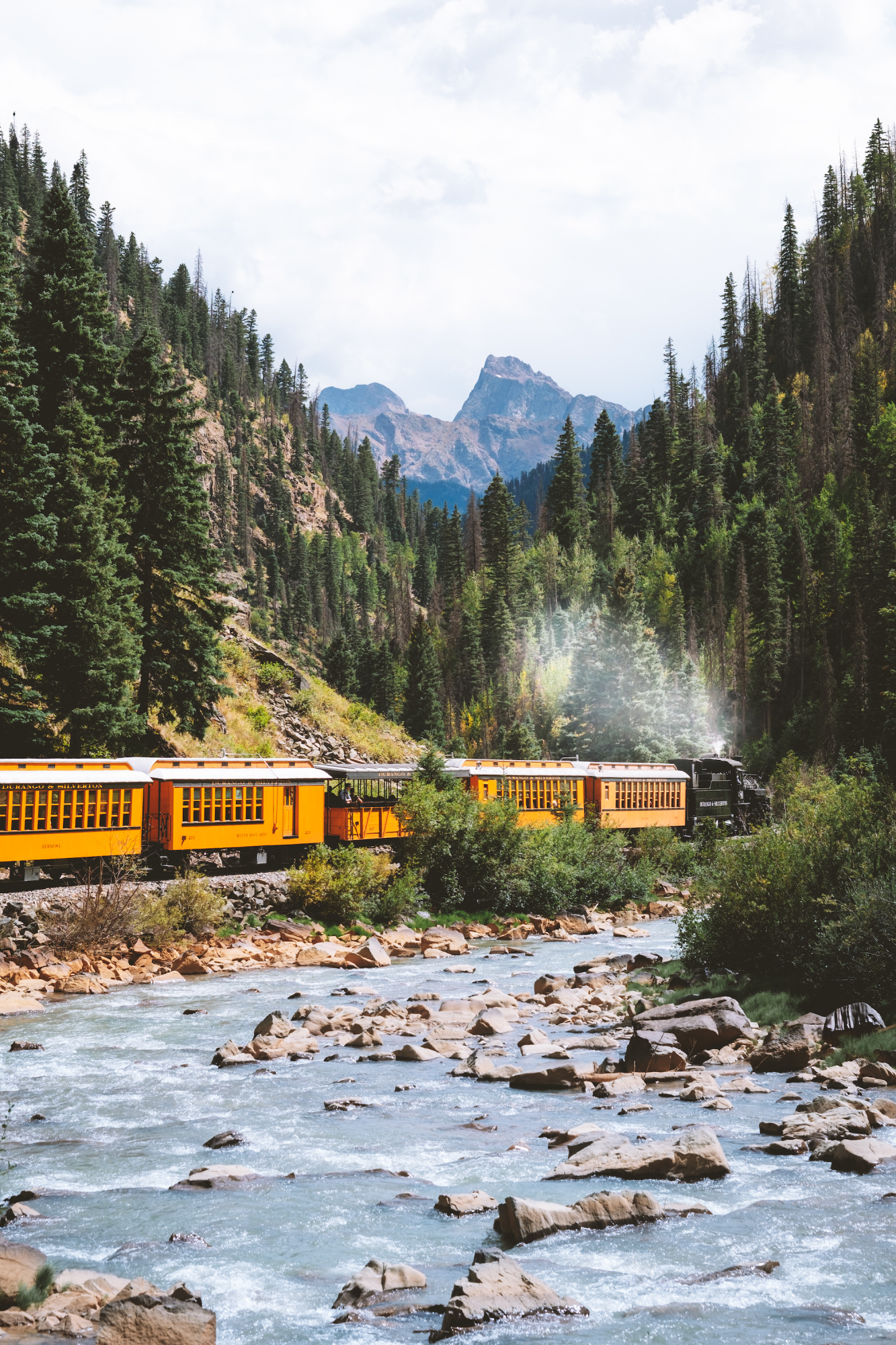 Dampfzug mit gelben Waggons fährt entlang eines Gebirgsflusses durch Durango, Colorado