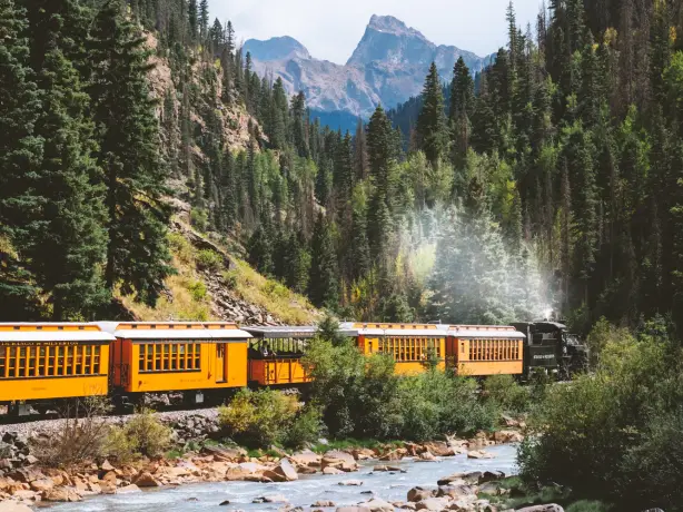 Dampfzug mit gelben Waggons fährt entlang eines Gebirgsflusses durch Durango, Colorado