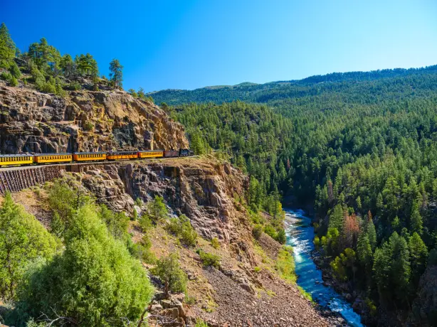Eine gelbe Dampflock fährt an einer Klippe, einem Gebirgsfluss und einem Wald entlang in Durango, Colorado