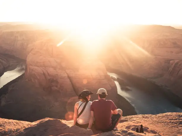 Ein Paar sitzt mit Blick auf Horseshoe Bend in Moab, Arizona, während die Sonne direkt in die Linse scheint.