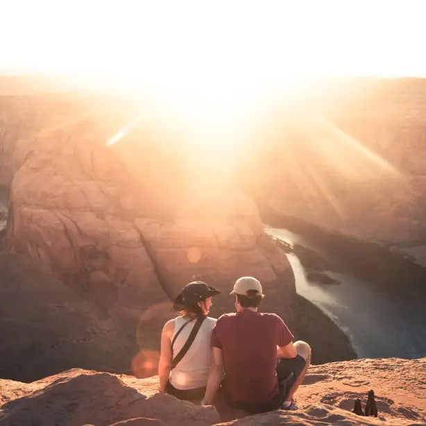 Ein Paar sitzt mit Blick auf Horseshoe Bend in Moab, Arizona, während die Sonne direkt in die Linse scheint.