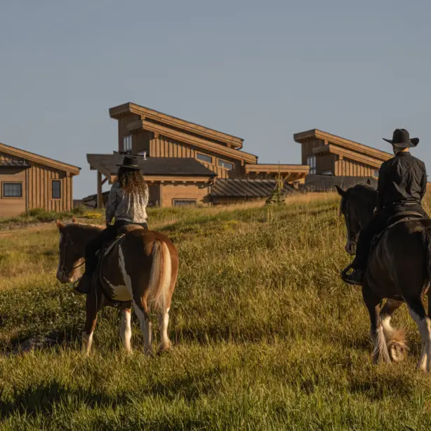 Twee ruiters met cowboyhoeden te paard die een met gras begroeide heuvel oversteken in de richting van een rustieke houten lodge in de buurt van Glacier National Park, Montana, VS, in het warme gouden licht van de middag.