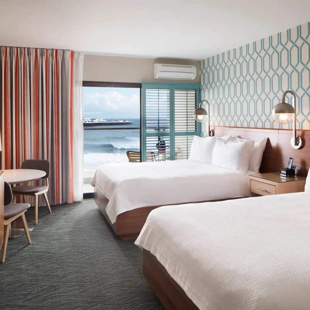 Helles Retro-Hotelzimmer in Santa Cruz, Kalifornien USA, mit zwei Queensize-Betten, blaugrüner geometrischer Tapete und einem Balkon mit Blick auf den Ozean und den Pier