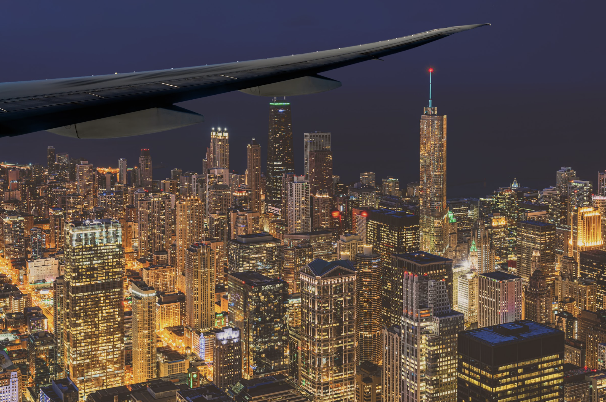 Luchtfoto van de verlichte skyline van Chicago bij nacht, genomen vanuit het raam van een vliegtuig, met de iconische wolkenkrabbers van de stad die onder de vleugel van het vliegtuig oplichten, VS