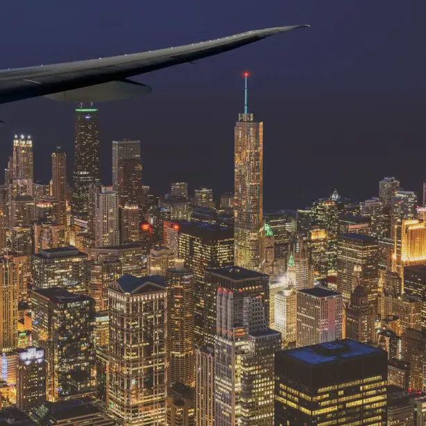 Luchtfoto van de verlichte skyline van Chicago bij nacht, genomen vanuit het raam van een vliegtuig, met de iconische wolkenkrabbers van de stad die onder de vleugel van het vliegtuig oplichten, VS