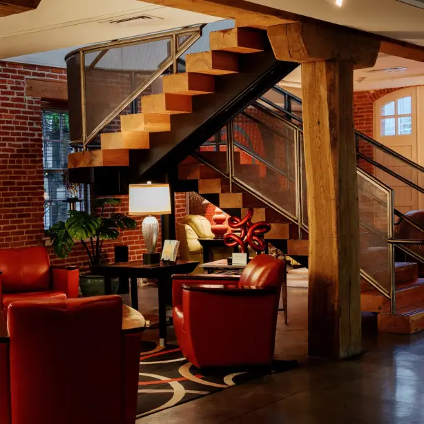 Stilvolle Boutique-Hotel-Lobby in Pennsylvania, USA, mit freiliegenden Backsteinwänden, schweren Holzbalken, roten Ledersesseln und einer skulpturalen schwebenden Treppe in einem historischen Gebäude