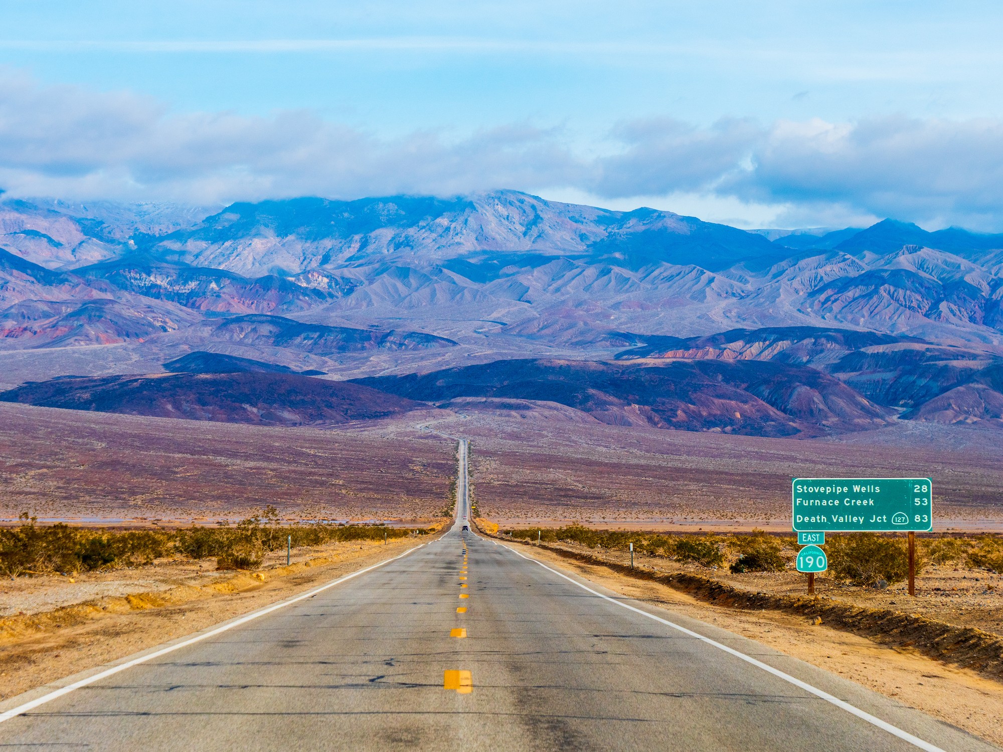 Vor uns erstreckt sich eine lange, offene Straße, während sich in der Ferne die Wüste und die Berge der Mojave-Wüste in Kalifornien abzeichnen