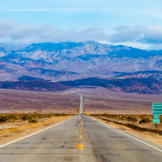 Vor uns erstreckt sich eine lange, offene Straße, während sich in der Ferne die Wüste und die Berge der Mojave-Wüste in Kalifornien abzeichnen
