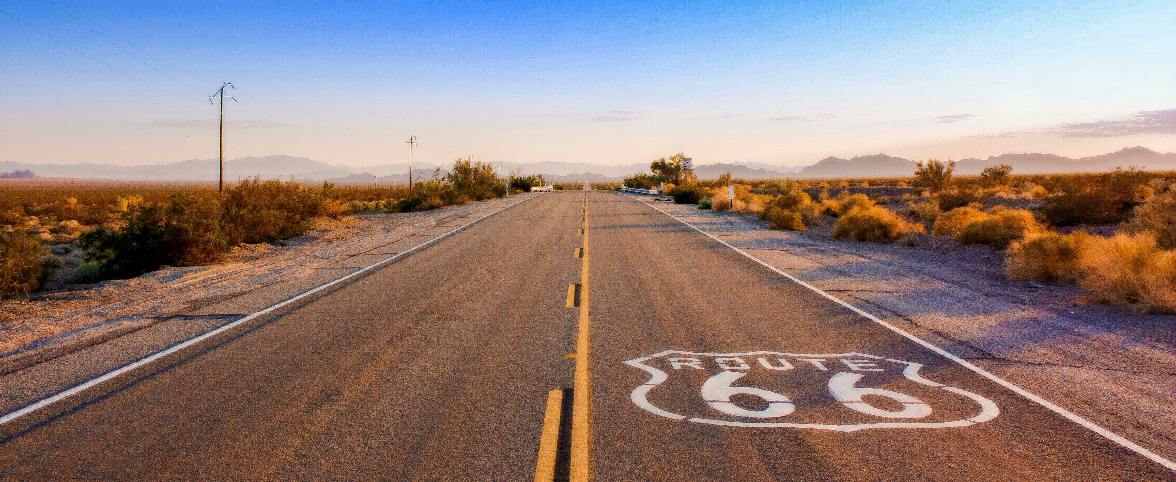 Eine leere Strecke der Route 66, die bei Sonnenaufgang durch die Wüste führt, mit langen Schatten und fernen Bergsilhouetten.